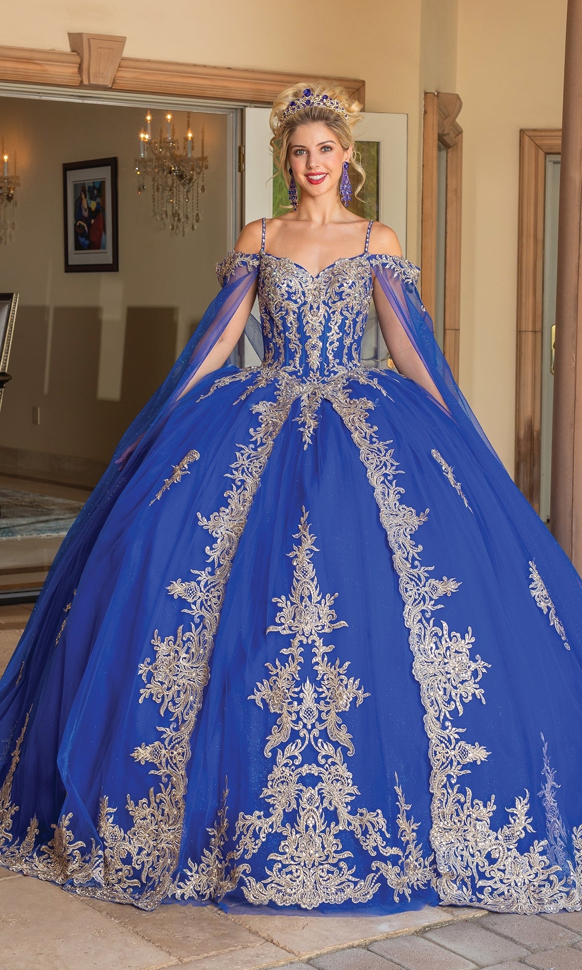 A woman poses gracefully indoors in the Quinceanera Dress 1768 By Dancing Queen—a royal blue off-the-shoulder ball gown with gold embroidery, matching accessories, and a tiara—standing near an open doorway.